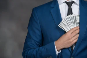 Torso of man wearing white dress shirt, black tie, and blue suit slipping fan of 14 $100 bills into jacket