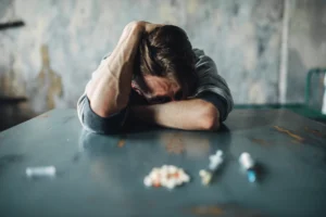 Pile of drugs, including orange and white tablets and two syringes, lying in front of man sitting head down at table
